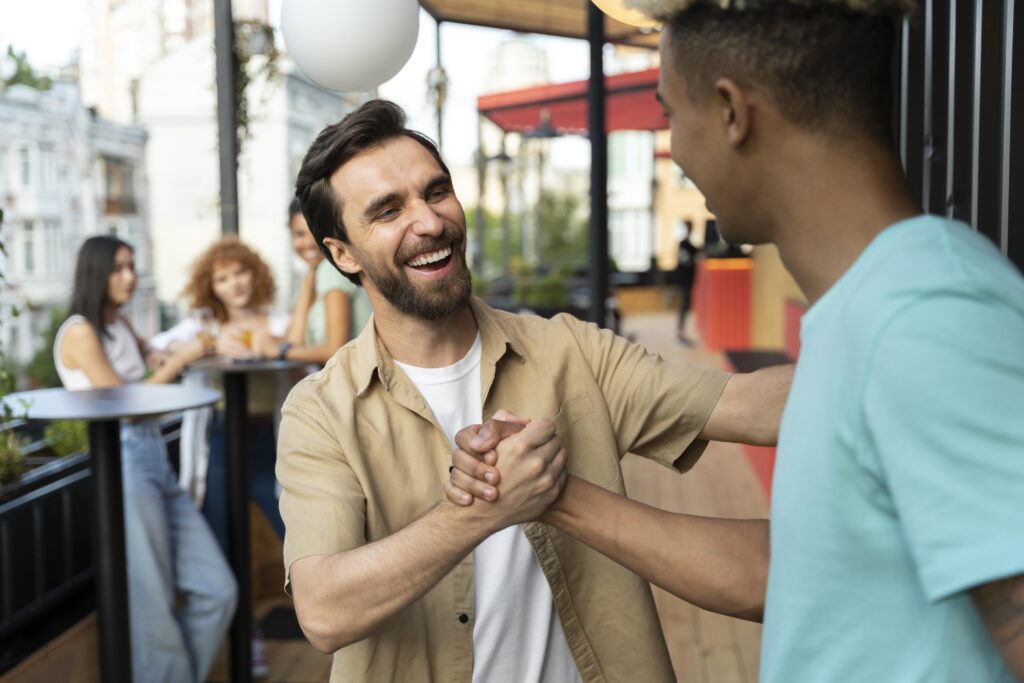 Dois homens compartilham um aperto de mão amigável, sorrindo e aproveitando o momento em um encontro social, com um grupo de pessoas ao fundo.