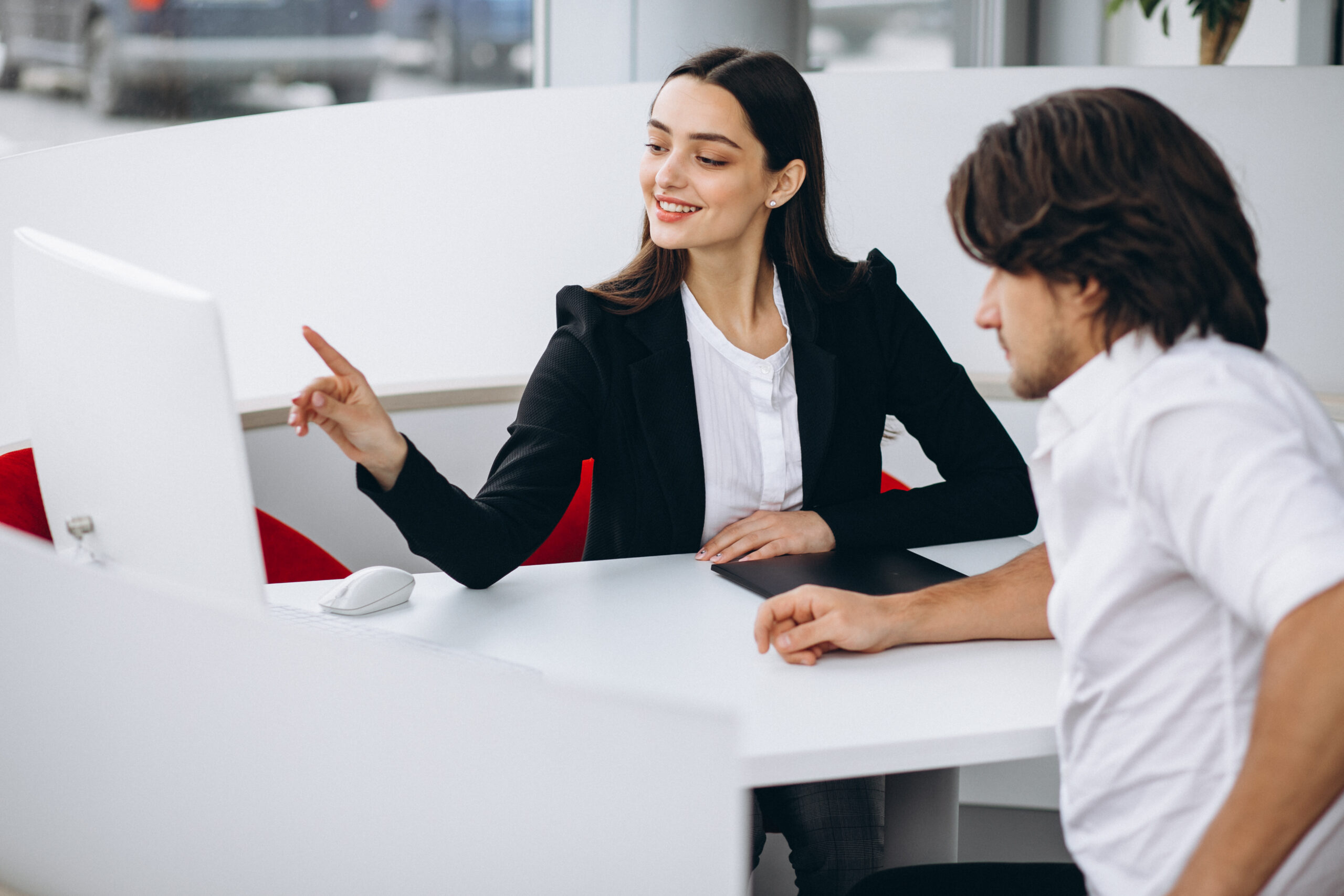 Uma mulher em um ambiente profissional aponta para a tela de um computador enquanto discute um projeto com um colega, ambos engajados na conversa.