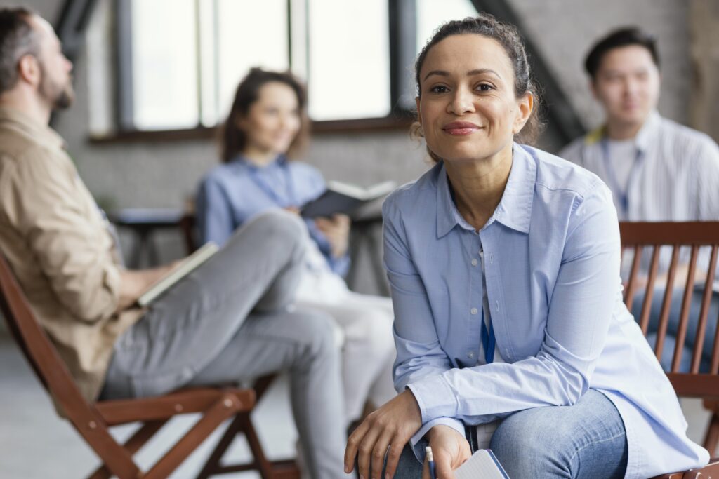 Mulher sorridente, usando camisa azul-clara e calça jeans, sentada e segurando um caderno, em um ambiente de reunião informal com outras três pessoas desfocadas ao fundo.