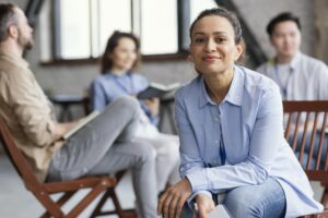 Mulher sorridente, usando camisa azul-clara e calça jeans, sentada e segurando um caderno, em um ambiente de reunião informal com outras três pessoas desfocadas ao fundo.