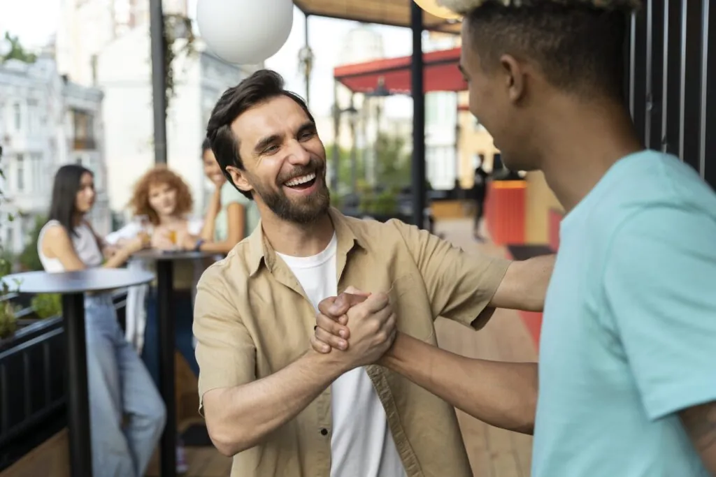 Dois homens compartilham um aperto de mão amigável, sorrindo e aproveitando o momento em um encontro social, com um grupo de pessoas ao fundo.