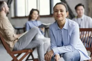 Mulher sorridente, usando camisa azul-clara e calça jeans, sentada e segurando um caderno, em um ambiente de reunião informal com outras três pessoas desfocadas ao fundo.